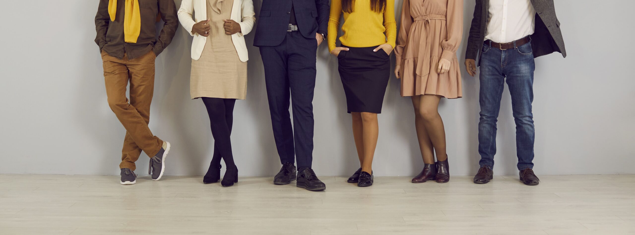 A group of six people stands against a light gray wall, shown only from the shoulders down. Their outfits are business casual, with earthy and vibrant tones.